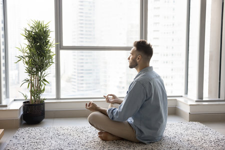 Peaceful focused young Latin yogi man meditating at home, keeping zen hands and meditating pose,...