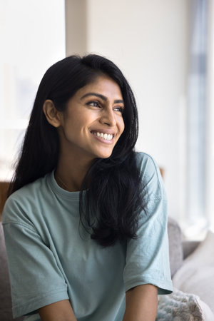 Positive attractive young 20s Indian woman in casual sitting on home sofa, looking away, thinking on...