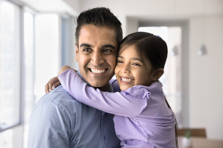 Happy loving 40s Indian dad holding adorable preschool daughter girl in arms, looking away with toothy smile, posing for home portrait. Happy father hugging sweet kid, enjoying fatherhoodの写真素材