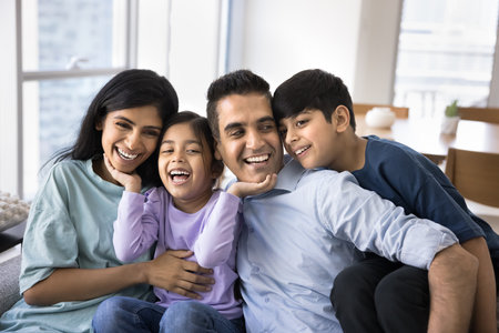 Happy excited Indian parents and two kids having fun at home, sitting close on couch, hugging,...