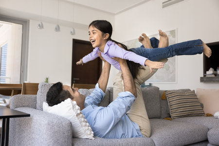 Cheerful little Indian kid and happy dad playing airplane together, laughing, having fun, enjoying activity in living room. Strong daddy holding excited little girl with open flying hands in armsの写真素材