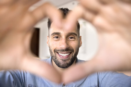 Happy handsome young Hispanic man looking at camera with face and toothy smile in finger heart...