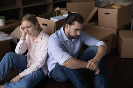 Upset unhappy young couple moving out from home, sitting at pile pf cardboard boxes, thinking over...