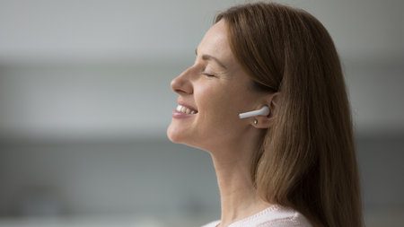 Side profile face view peaceful pretty woman enjoy music through wireless earphones standing indoors...