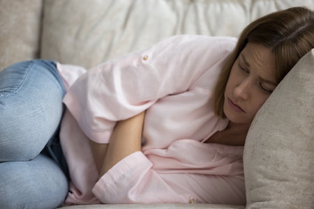 Close up young woman feels unwell, looks unhealthy, lying on sofa hugs her stomach suffers from menstrual pain period or stomachache, lonely unhappy female think of life concerns, goes through divorceの写真素材