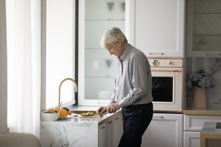 Older retired homeowner man cooking salad in modern kitchen, chopping fresh vegetables on board, preparing organic meal for dinner from natural ingredients. Healthy eating, elderly agの写真素材
