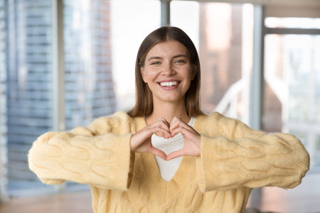 Cheerful beautiful young woman in warm sweater showing hand heart, symbol of love, health, gratitude, charity. Blogger girl thanking audience, looking at camera with toothy smile. Head shot portraitの写真素材