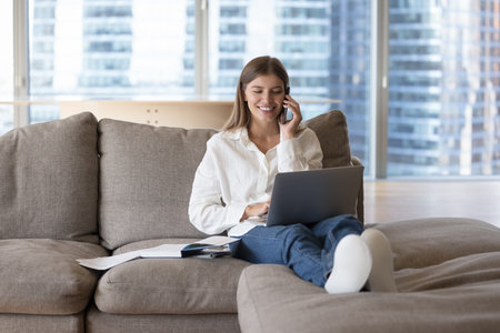 Happy young freelancer girl answering mobile phone call on home couch, speaking to customer on telephone, typing on laptop computer, using online app, enjoying job Internet communication, smilingの写真素材