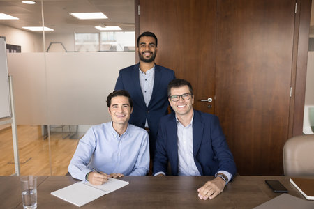 Happy successful diverse male business team posing together, sitting and standing at meeting table,...