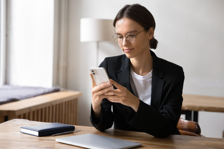 Attractive businesslady sit at workplace desk with smartphone, using mobile banking or financial management tools, planning, organizing workday, solve work-related matters efficiently via applicationの写真素材