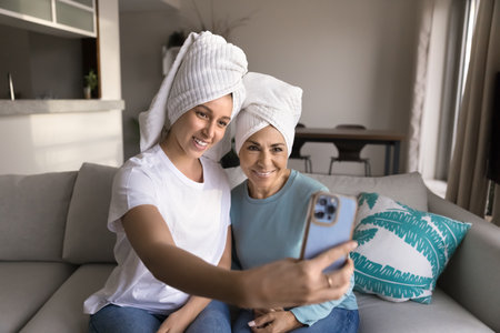Happy beautiful young Latin daughter and mature mom in head towels taking selfie on smartphone, having fun, enjoying beauty care activities at home, smiling, laughing at mobile phoneの写真素材