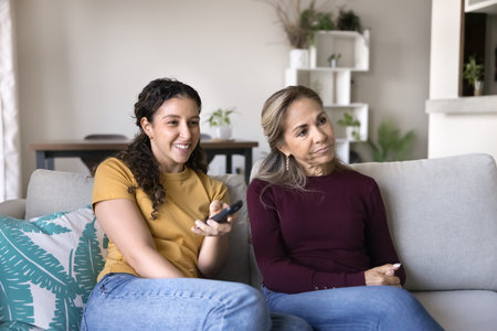 Cheerful young adult Hispanic daughter girl and annoyed bored mom watching comedy series, funny show...
