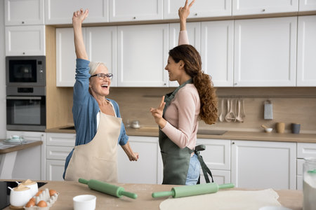 Lively older woman and millennial daughter wear aprons dancing in kitchen, moving to favourite music with arms raised, feel happiness, looking carefree, while preparing home-made holiday cake at homeの写真素材