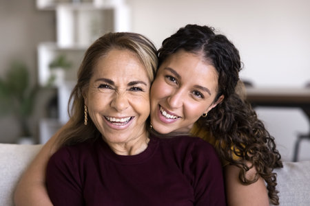 Cheerful beautiful young daughter woman embracing happy mature Latin mother with love, care, gratitude, looking at camera, smiling, laughing, posing for family portraitの写真素材