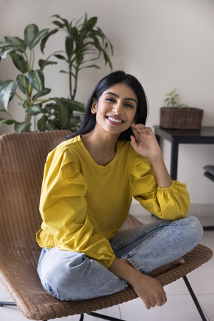Happy beautiful young Indian woman sitting on home lounge chair, looking at camera with toothy...