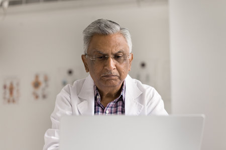 Focused older 65s male doctor dressed in white lab coat working on laptop, accessing and updating medical histories, and treatments to track progress, conducting virtual consultations with patientsの写真素材