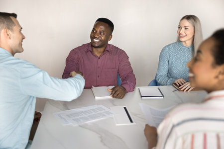 Diverse male business partners shaking hands at large office table shared with team, meeting for...