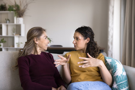 Positive mature Latin mom and young adult daughter woman sitting on home couch, talking, discussing family event, relationship, enjoying meeting, offline communicationの写真素材