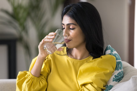 Peaceful satisfied young Indian woman drinking fresh pure water with closed eyes at home, holding...