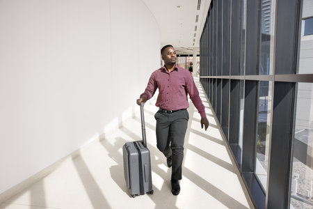 Young African American businessman walking in hurry to airport flight gate, wheeling suitcase in corridor, looking at window, enjoying business trip, travel abroad, tourism, voyageの写真素材