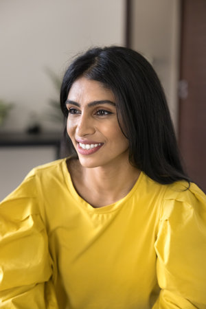 Positive dreamy beautiful Indian woman in yellow cloth looking away, smiling, thinking, daydreaming. Happy young female model with long black hair posing for casual vertical portrait at homeの写真素材