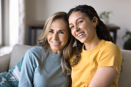 Happy Latin young adult daughter woman and mature mom sitting close on couch, smiling, laughing, posing for home family portrait, looking away, enjoying friendship, affectionate relationshipの写真素材