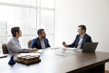 Diverse team of male business leaders in formal suits talking at table in meeting room, discussing teamwork, collaboration, partnership. Board of directors, stakeholders wide shotの写真素材