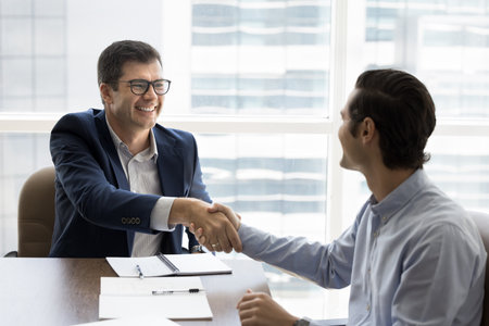 Happy young businessman giving handshake to financial consultant, legal advisor after consultation. Two male business partners shaking hands over meeting table, closing deal, getting agreementの写真素材