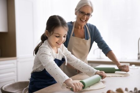 Cute girl flattening dough with rolling-pin helps with cooking process to 60s granny, wear aprons preparing holiday family recipe pie in modern kitchen. Multi-generational relatives joint activityの写真素材