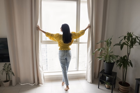 Beautiful black haired young woman standing at large window in stylish cozy apartment, opening curtains, enjoying day light, city view from higher floor. Wide full length shotの写真素材