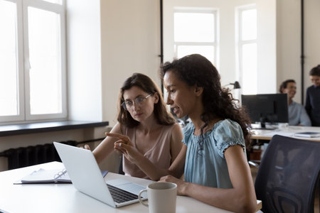 Female colleagues sit at desk use laptop talk about project startup ideas, share thoughts, solve business issues, make research, discuss growth strategy, analyze revenue at workplace. Teamwork conceptの写真素材