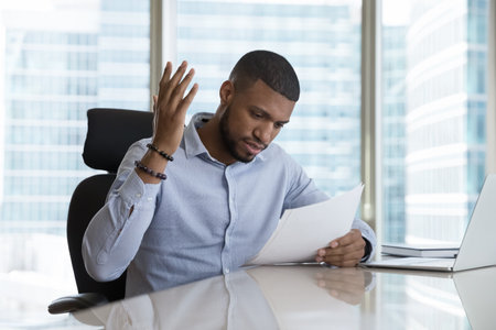 Frustrated African man read bad news in postal letter or paper document sit at desk in modern skyscraper office, get negative response, dismissal notice, high bills, huge taxes, bank debt notificationの写真素材