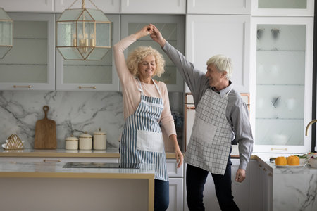 Attractive mature wife and older husband in aprons holding hands swirling having fun distracted from cooking in fashionable kitchen, moving to music, dancing together at home feel carefree look happyの写真素材