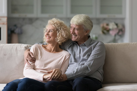Elderly couple relaxing on sofa staring into distance. Serene attractive older wife, grey haired husband smile lookaside feeling carefree, enjoy calm retired life. Well-being family, happy marriageの写真素材