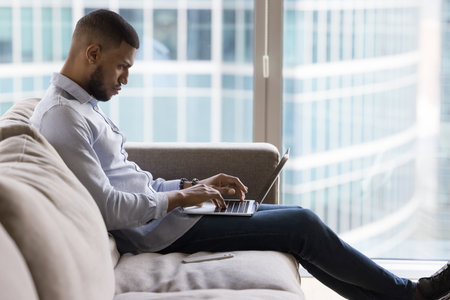 Focused African man buy goods, typing e-mail, work or study on laptop, sits on couch in modern flat. E-commerce client makes order, purchasing electronic services, doing telework at home use internetの写真素材