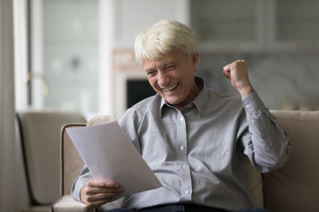 Overjoyed senior man sitting on sofa with papers, reading letter feels excited by good fantastic...