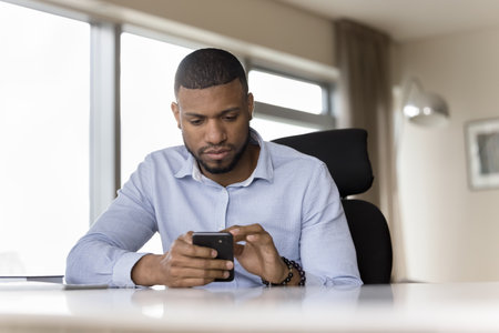 Successful serious millennial African businessman using smartphone seated at workplace in modern...