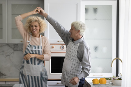 Pretty older spouses, couple in love in aprons dance in kitchen, holding hands swirling moving to music, feel happy, enjoy carefree retired life and cookery together at home. Romantic relations, loveの写真素材