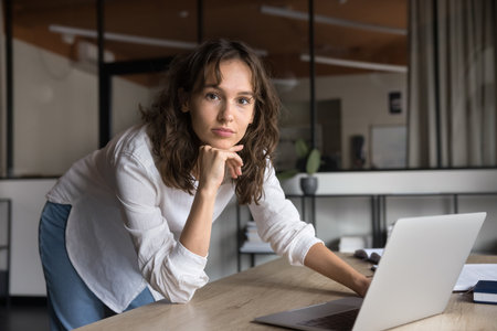 Young woman working on laptop, posing for camera with pensive expression, engaged in problem-solving or decision-making, preparing presentation, analyze data. Project management, consulting, workflowの写真素材