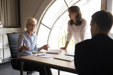 Group of young employees listening aged boss of company, gathered together at conference table for financial planning or budget discussion, problem-solving, brainstorming new ideas and setting goalsの写真素材