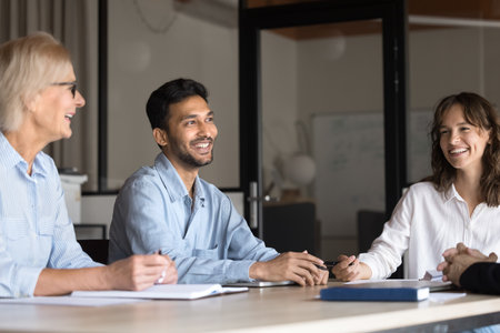 Happy teammates lead informal talk seated at conference table. Multiethnic businesspeople or stakeholders take part in meeting in company office, laughing, joking, feel satisfied with briefing outcomeの写真素材