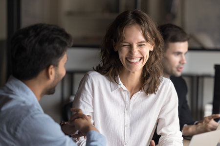 Happy woman employee laughing while talking with Indian man teammate sit at desk enjoy friendly...