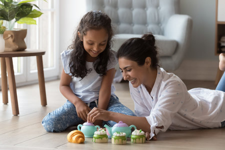 Happy engaged mom and cute little daughter pretending drinking tea from plastic dish with toy baked sweet food, playing coffee shop role game, sitting, resting on heating floor at home, having funの写真素材