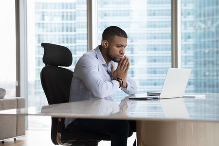 Serious young African business man looking at laptop screen, sitting at urban office work table...