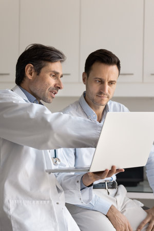 Serious middle aged practitioner doctor showing medical report to patient, pointing at laptop screen, explaining test results, using modern technology on consultationの写真素材