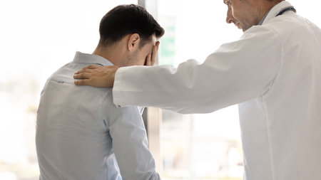 Compassionate male doctor telling hard serious diagnosis to patient after medical checkup, touching shoulder of stressed shocked man covering face in supportive, sympathetic gesture. Banner shotの写真素材