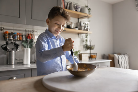 Happy positive boy kid stirring flakes and milk in bowl, eating at home, having breakfast in kitchen, holding spoon, enjoying tasty delicious food, preparing healthy cereal by himself, smilingの写真素材