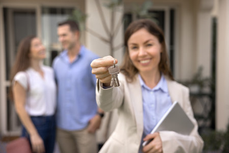 Happy successful female real estate agent offering key, showing bunch at camera, posing for professional realtor portrait with rental accommodation and couple of renters, buyers in blurred backgroundの写真素材