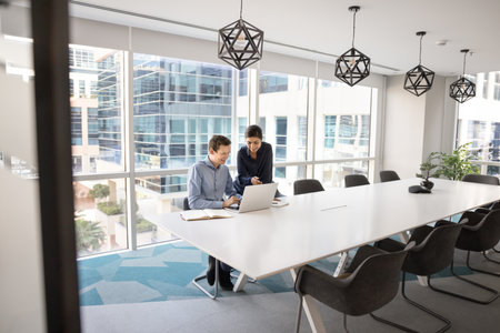Multiethnic couple of positive managers watching online content on laptop in office meeting room, sitting at large conference table alone, using online communication for teamwork. Candid wide shotの写真素材
