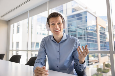 Happy young business professional man wearing headphones, speaking at camera, talking on online...